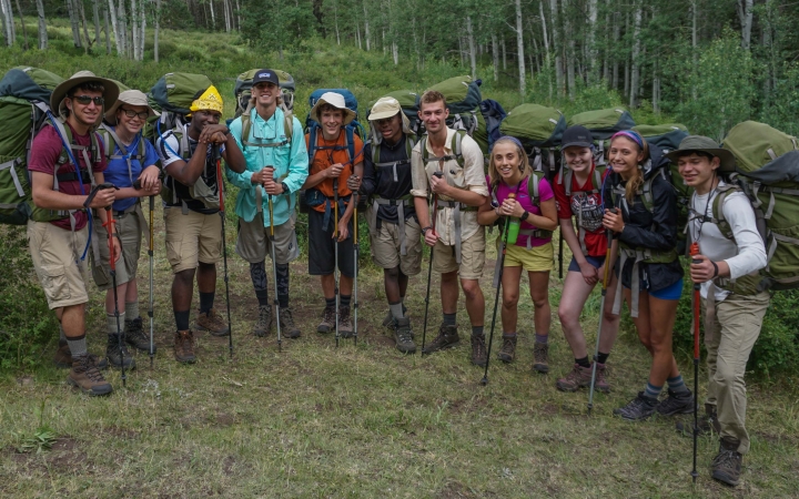 A group of students wearing backpacks smile for a photo in a green meadow 